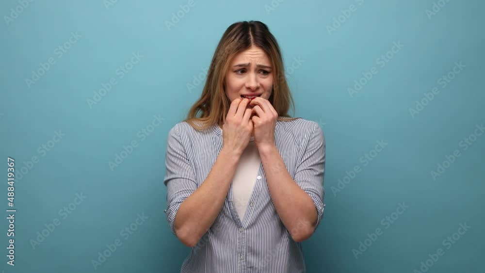 Troubles and worries. Portrait of nervous woman biting nails, terrified about problems, suffering phobia, anxiety disorder, wearing striped shirt. Indoor studio shot isolated on blue background. Troubles and worries. Portrait of nervous woman biting nails, terrified about problems, suffering phobia, anxiety disorder, wearing striped shirt. Indoor studio shot isolated on blue background.