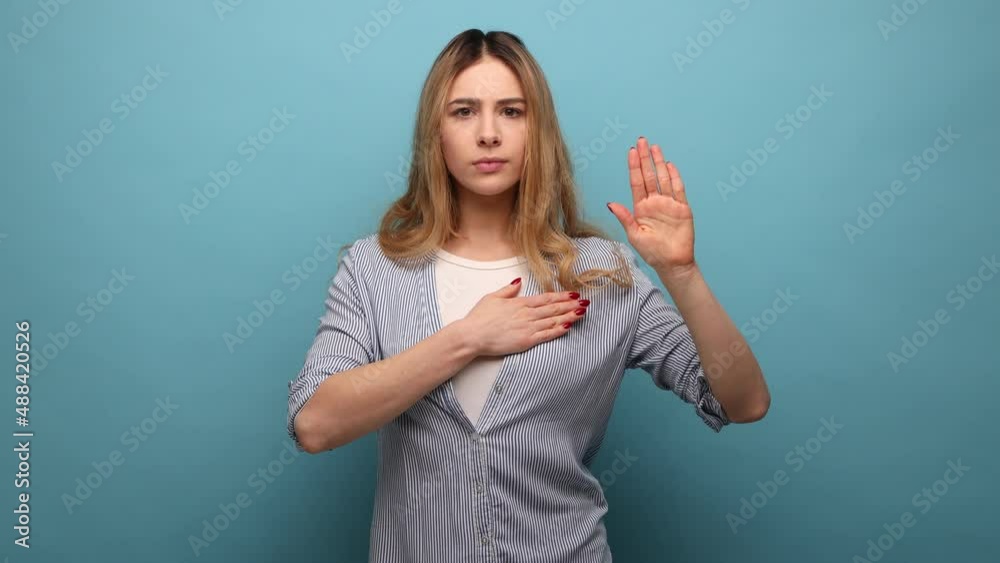Portrait of very responsible and honest woman giving promise, making solemn vow in ceremonial tradition with raised hand, wearing striped shirt. Indoor studio shot isolated on blue background.