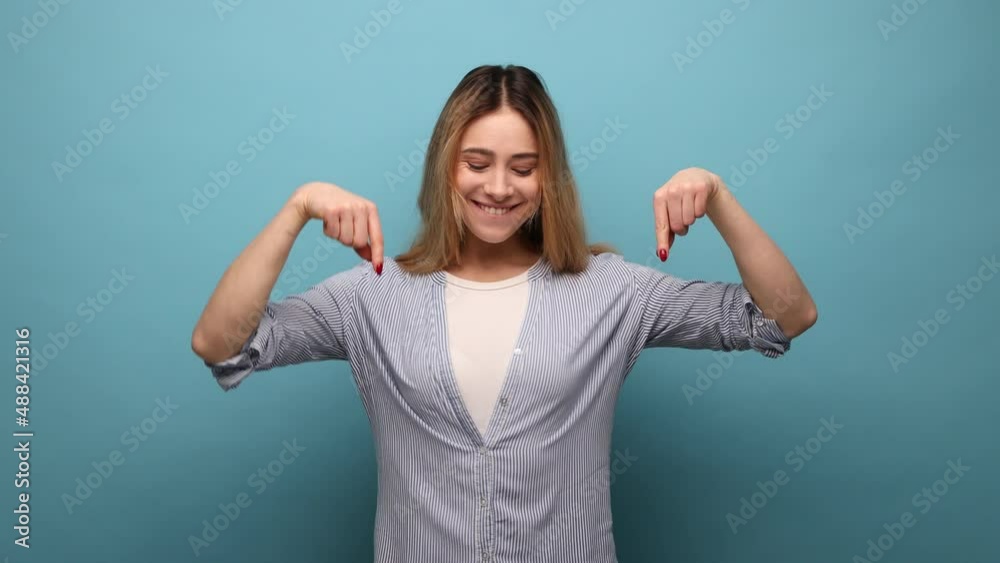 Portrait of positive wavy haired woman pointing down place for commercial idea, looking at camera with toothy smile, wearing striped shirt. Indoor studio shot isolated on blue background.