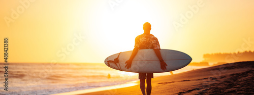 Silhouette of surfer in hawaiian t-shirt walking with surf board on the beach at sunset, panoramic