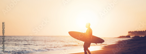 Surfer in hawaiian t-shirt with surf board on the beach at sunset, walking to the ocean, panoramic