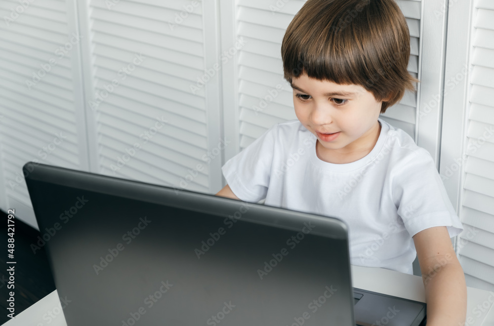 5 year boy sit at the table, uses laptop, looking at the screen. Child is doing homework lesson, playing video games, studying IT development, watching cartoon. Home distance online education concept