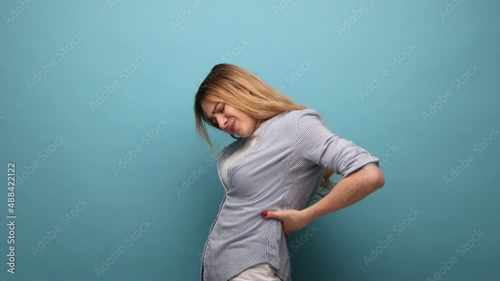 Portrait of frustrated anxious unhealthy woman holding hands on lower back, kidney injury, backache, feeling pain, wearing striped shirt. Indoor studio shot isolated on blue background.