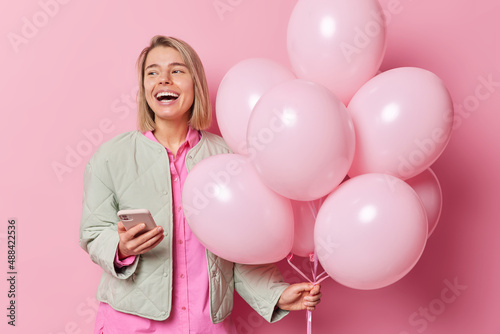 Glad fair haired woman uses mobile phone sends text messages comes on party enjoys celebration holds bunch of inflated balloons looks away happily wears jacket isolated over pink background.