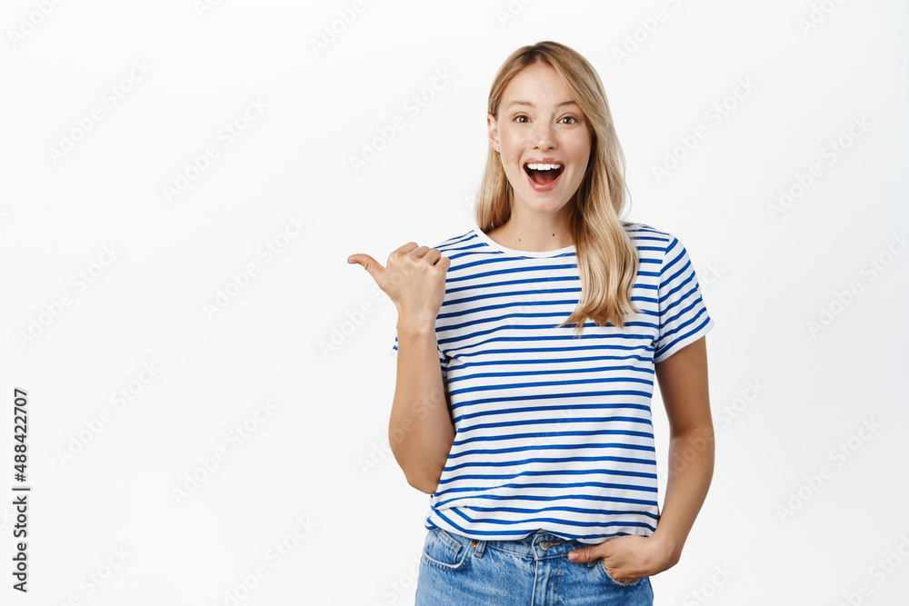 Happy smiling blond woman pointing finger left, showing announcement or banner, announcing event, standing in tshirt over white background Happy smiling blond woman pointing finger left, showing announcement or banner, announcing event, standing in tshirt over white background