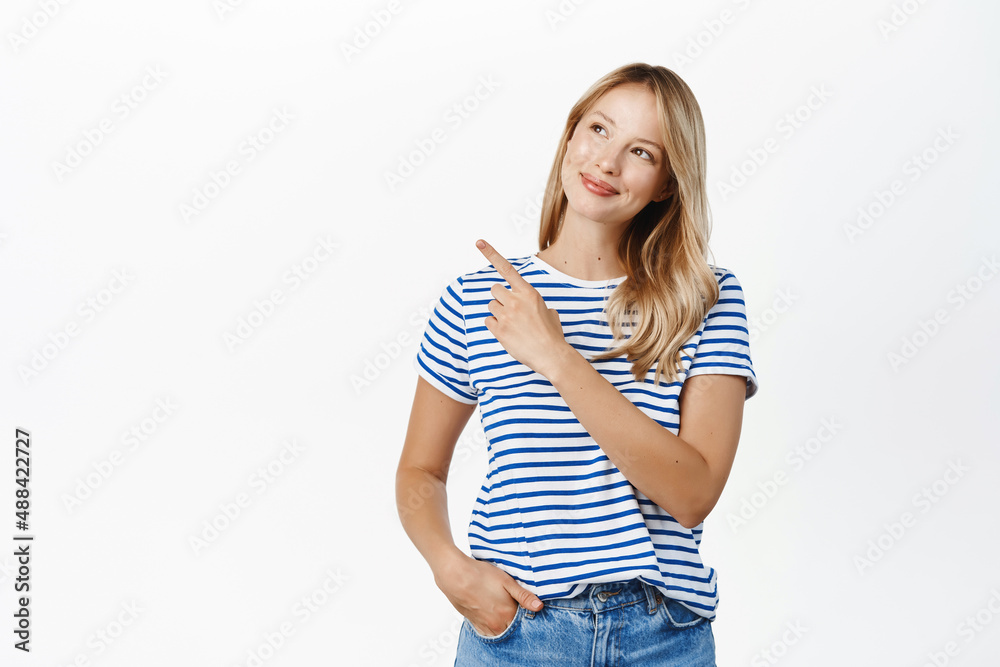 Portrait of young woman looks thoughtful and points at upper left empty space, promo banner on top, thinking or choosing in store, standing over white background Portrait of young woman looks thoughtful and points at upper left empty space, promo banner on top, thinking or choosing in store, standing over white background