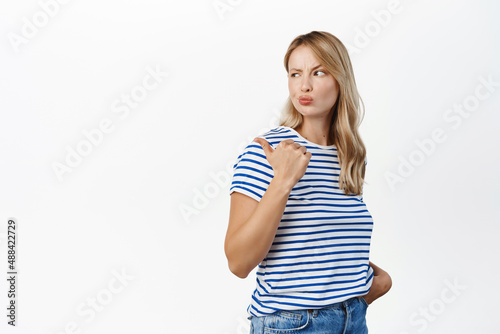 Image of modern young woman with blond hair, looks skeptical and doubtful, pointing finger left at suspicious sale, unsure about product, white background