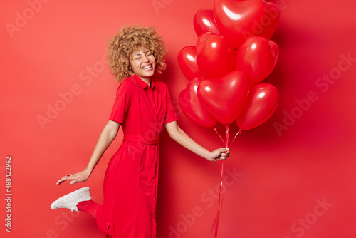 Happy energetic curly haired woman has fun dances and raises leg expresses positive emotions holds bunch of inflated heart shaped helium balloons comes on party isolated over vivid red background.