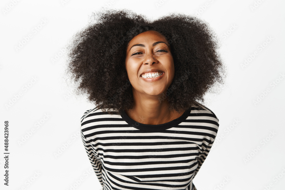 Beautiful african american woman shows her white healthy smile, smiling and laughing, looking happy, standing in striped blouse over white background