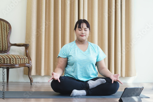 Young woman sitting on yoga mat with Technology tablet online, during meditation practicing yoga in sukhasana exercise at yoga studio. Selection focusing on hand foreground with copy space.