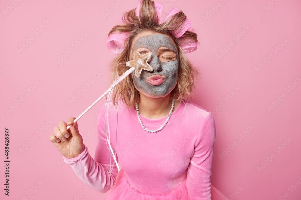 Horizontal shot of female model keeps lips rounded covers eye with magic wand applies nourishing clay mask to reduce fine lines makes hairstyle wears festive dress and necklace isolated on pink wall Horizontal shot of female model keeps lips rounded covers eye with magic wand applies nourishing clay mask to reduce fine lines makes hairstyle wears festive dress and necklace isolated on pink wall