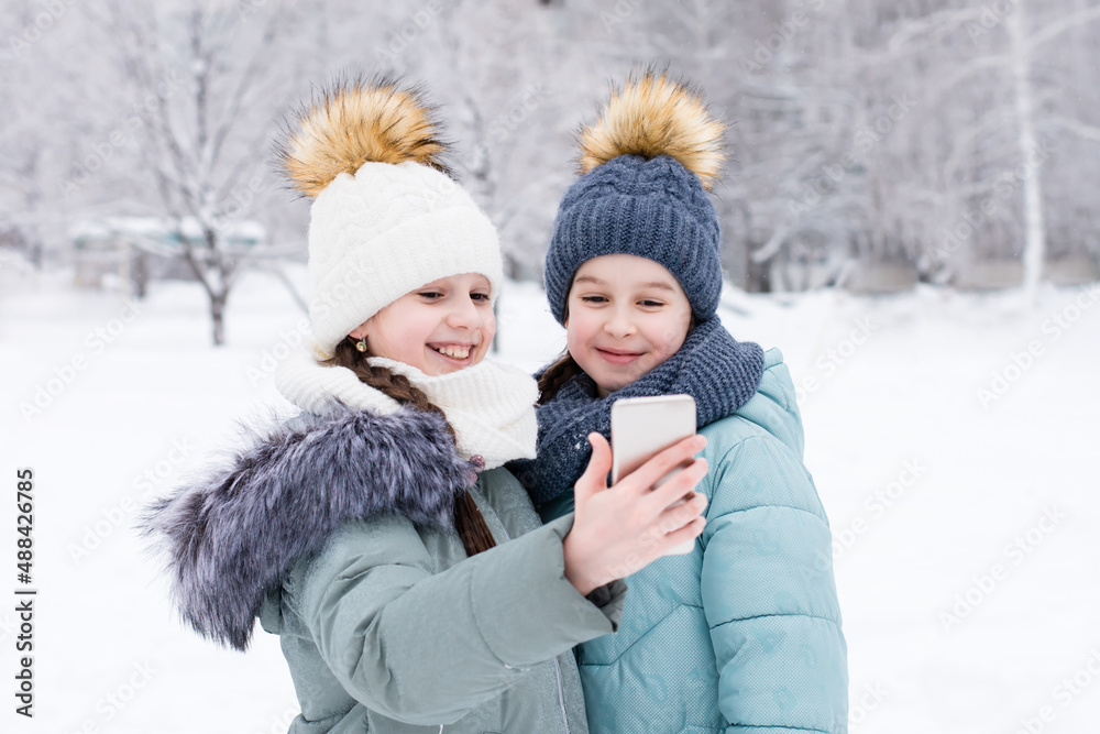 Two smiling girls in warm clothes make a video call on a smartphone in a snowy winter park. Lifestyle use of technology