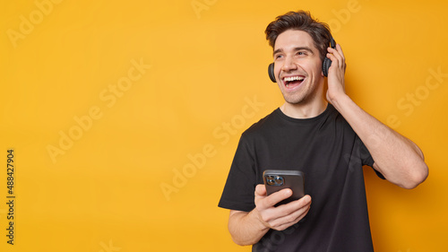Horizontal shot of happy adult man dressed in casual black t shirt listens music via headphones enjoys favorite audio track isolated over yellow background blank copy space for your promotion