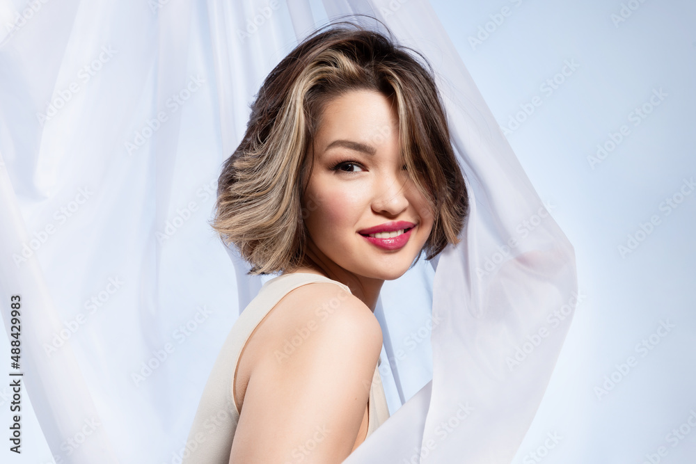Friendly beautiful young woman with short haircut, perfect fresh makeup posing near white waving cloth. Flirtatious playful woman looking at camera on light curtain background. Friendly beautiful young woman with short haircut, perfect fresh makeup posing near white waving cloth. Flirtatious playful woman looking at camera on light curtain background.