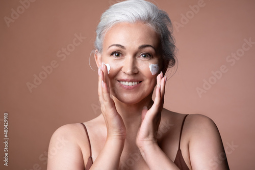 Happy senior female smiling apply antiaging rejuvenating cosmetic face cream. Middle aged woman care healthy skin applying beauty cosmetics moisturizing product. Aging and skincare concept.