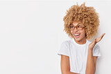 Horizontal shot of crazy curly woman reacts emotionally on something keeps palm raised smiles happily wears casual t shirt and spectacles poses against white background copy space for your promo