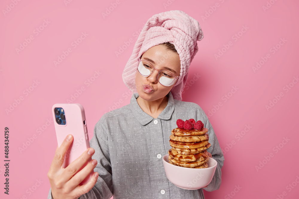 Lovely young woman makes selfie tilts head keeps lips rounded poses at smartphone camera poses with prepared pancakes with syrup and raspberries wears pajama towel wrapped on head. Happy awakening Lovely young woman makes selfie tilts head keeps lips rounded poses at smartphone camera poses with prepared pancakes with syrup and raspberries wears pajama towel wrapped on head. Happy awakening