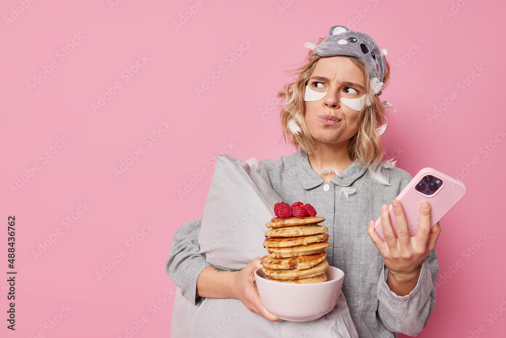 Unhappy annoyed woman looks angrily away makes plannings in mind uses mobile phone for surfing internet holds bowl of pancakes with raspberries applies beauty patches holds pillow stands indoor Unhappy annoyed woman looks angrily away makes plannings in mind uses mobile phone for surfing internet holds bowl of pancakes with raspberries applies beauty patches holds pillow stands indoor