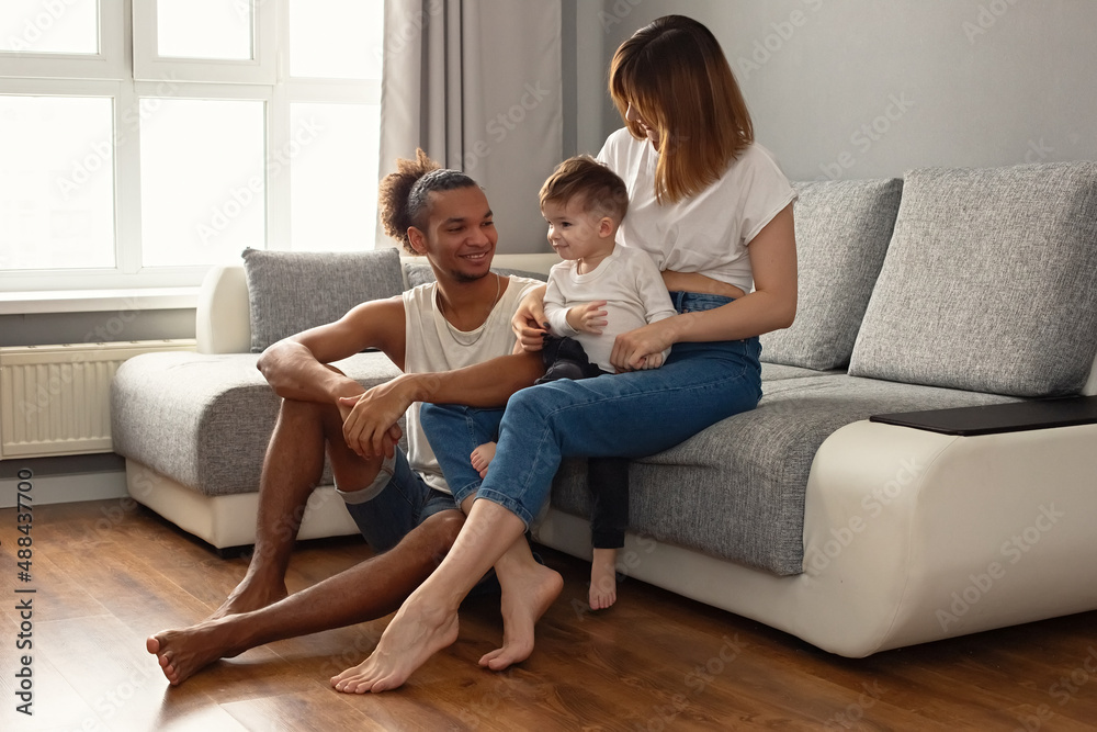 Happy, modern, multiracial family with a little boy in cozy home clothes sit on a gray sofa near a large window, smiling