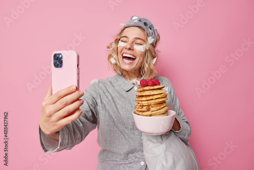 Happy positive young woman maes photo via smartphone laughs gladfully poses with tasty pancakes dressed in nightwear feels very glad enjoys weekend and lazy day spends it at home poses indoor photo