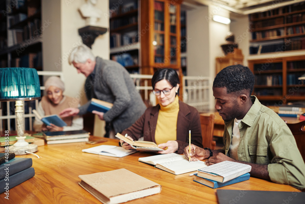 Diverse group of people sitting in row at table in college library and studying, focus on young black man reading book in foreground Diverse group of people sitting in row at table in college library and studying, focus on young black man reading book in foreground