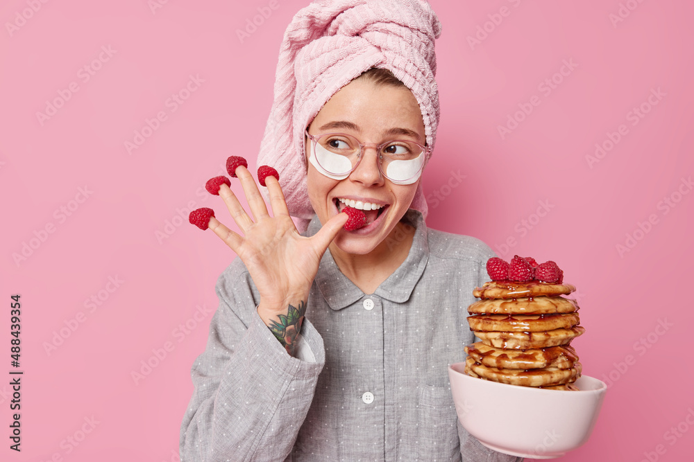 Pleased young woman has raspberries on fingers poses with delicious pancakes indoor wears pajama and wrapped towel on head applies beauty patches enjoys good morning isolated on pink studio wall Pleased young woman has raspberries on fingers poses with delicious pancakes indoor wears pajama and wrapped towel on head applies beauty patches enjoys good morning isolated on pink studio wall
