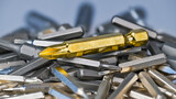 Big golden cross screwdriver bit on silvery bits heap of various sizes and types. Close-up of slot or cruciform screw drives with hex shank. Pile of replaceable metal tools on blurry white background.