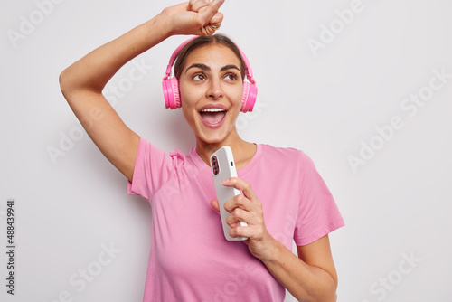 Energetic optimistic woman dances carefree sings favorite song enjoys playlist uses modern smartphone and stereo headphones dressed in pimk t shirt isolated over white background. Enternainment photo