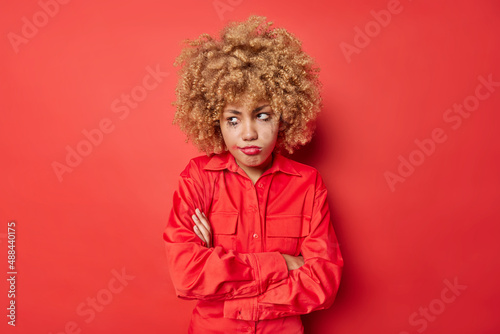 Displeased offended youg woman keeps arms folded looks unhappily away has hurt feelings has leaked makeup wears shirt isolated over red background. Moody gloomy female model feels depressed.