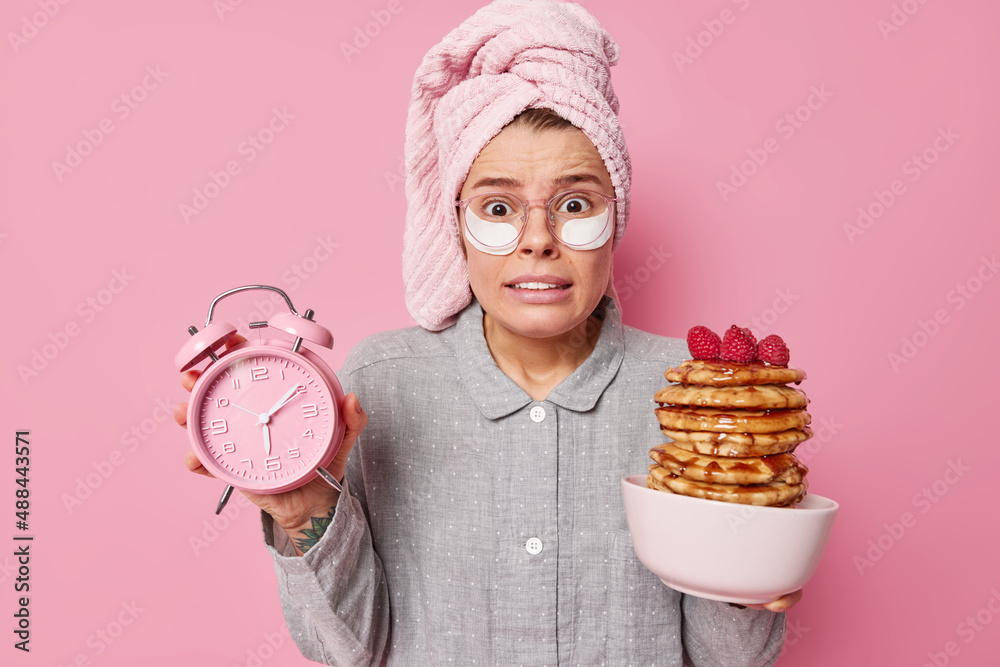 Embarrassed worried woman being in hurry holds alarm clock and delicious pancakess with syrup and raspberries wears comfortable pajama wrapped towe and big transparent glasses applies patches Embarrassed worried woman being in hurry holds alarm clock and delicious pancakess with syrup and raspberries wears comfortable pajama wrapped towe and big transparent glasses applies patches