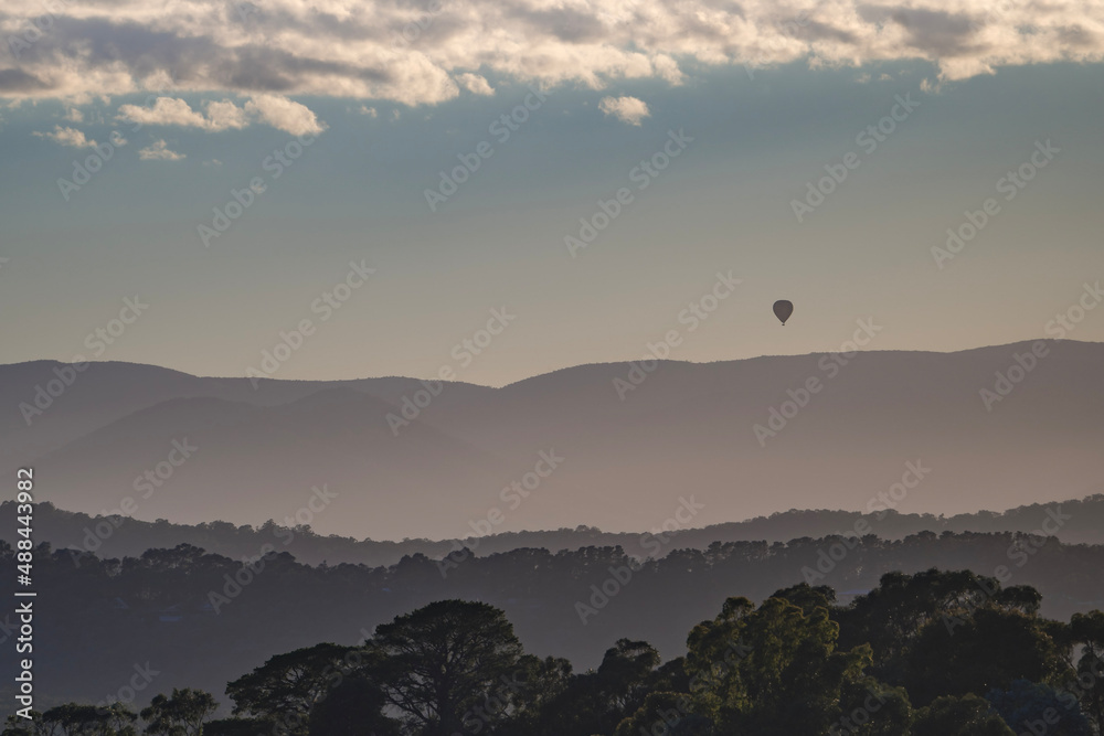 Single hot air balloon seen in silhouette floating in the distance over misty layers of mountains 