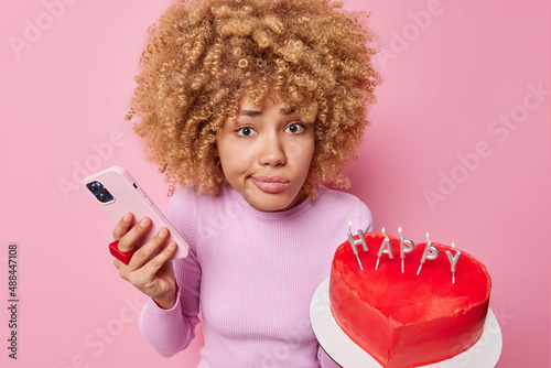 Indignant puzzled curly haired woman holds mobile phone and heart shaped cake prepares for celebration of special occasion waits forr guests poses against pink background. Happy birthday concept photo