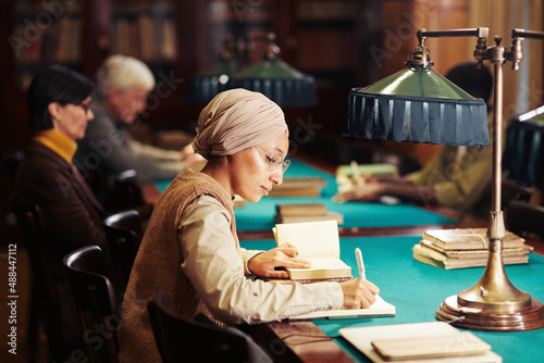 Side view portrait of young Muslim woman studying in classic library interior, copy space