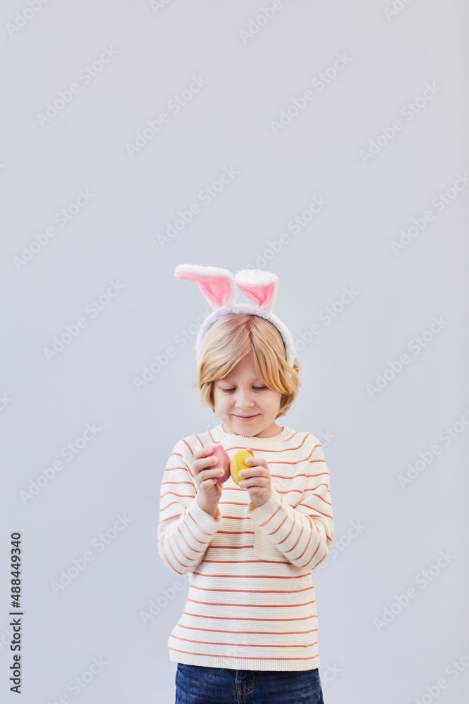 Vertical waist up portrait of cute blonde boy wearing bunny ears and playing with Easter eggs against neutral white background, copy space Vertical waist up portrait of cute blonde boy wearing bunny ears and playing with Easter eggs against neutral white background, copy space