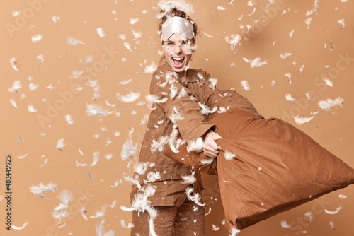 Positive funny young woman fights with pillow enjoys weeekend has happy expression smiles gladfully exclaims from joy wears sleepwear poses against brown background with feathers flying in air photo