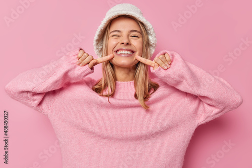 Look at my shining smile. Positive fair haired European woman points at her white teeth smiles broadly wears fashionable panama and casual jumper isolated over pink background has happy mood photo