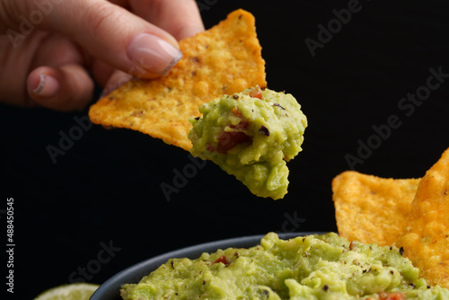 Hand with tasty tortilla chips or nachos with fresh homemade guacamole dip sauce on black background photo