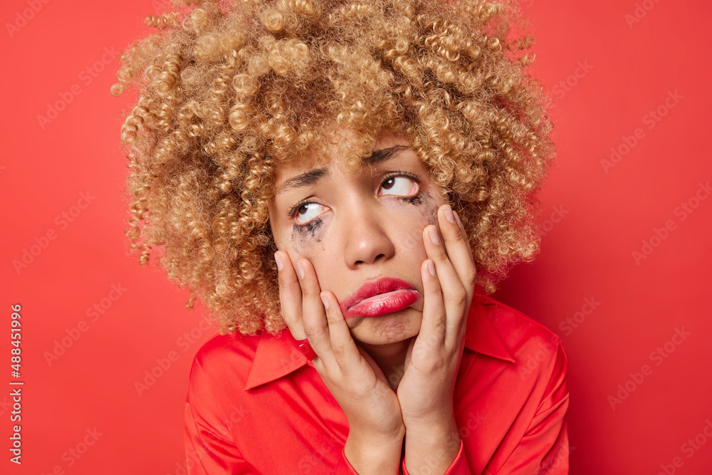 Upset crying woman with curly bushy hair has frustrated expression keeps hands on face looks with despair has broken heart feels hurt isolated over red background. Negative human emotions concept Upset crying woman with curly bushy hair has frustrated expression keeps hands on face looks with despair has broken heart feels hurt isolated over red background. Negative human emotions concept