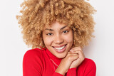 Portrait of good looking cheerful young woman with curly bushy hair smiles toothily keeps hands near face wears red turtleneck isolated over white background. Positive human emotions concept
