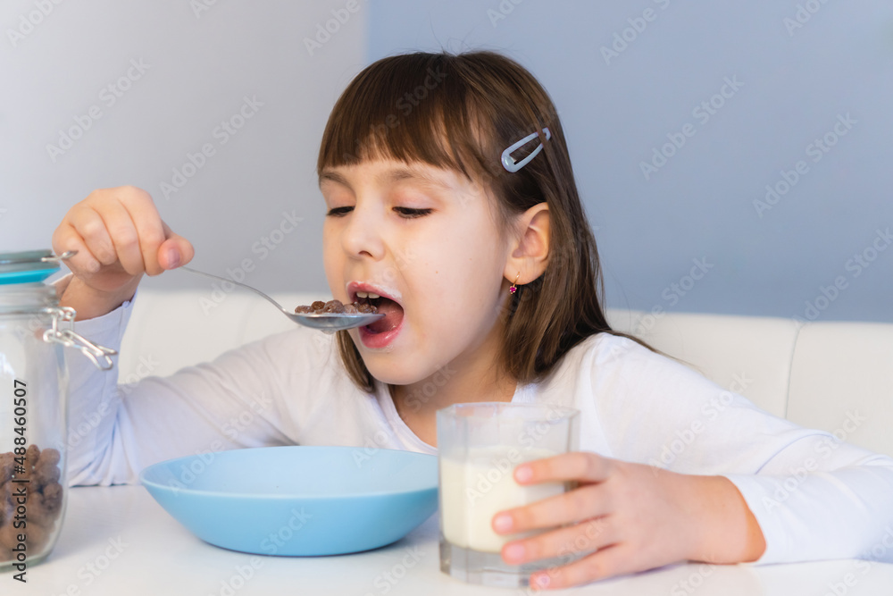 Little girl eats chocolate corn balls with milk sitting at white table. Child have breakfast Little girl eats chocolate corn balls with milk sitting at white table. Child have breakfast