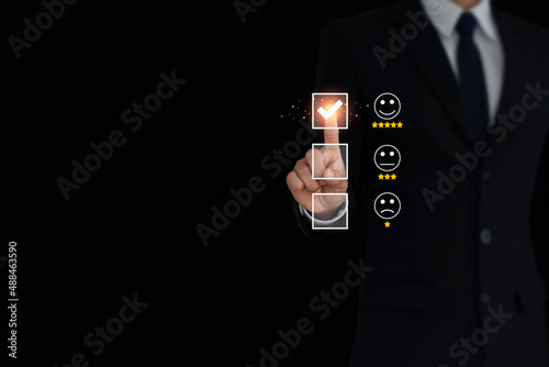 A businessman wearing black suit standing on black background presses a smiley face emoticon and select five star on a virtual touch screen great customer service with copy space.