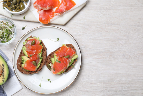 Delicious sandwiches with salmon, avocado and capers on white wooden table, flat lay. Space for text