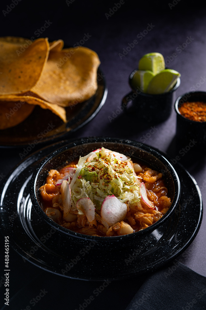 Traditional mexican food. Red pozole soup with chicken accompanied with avocado, lettuce, onion, radish, lemon, chili and crispy corn tortillas also known as tostadas on a black wooden background. Traditional mexican food. Red pozole soup with chicken accompanied with avocado, lettuce, onion, radish, lemon, chili and crispy corn tortillas also known as tostadas on a black wooden background.