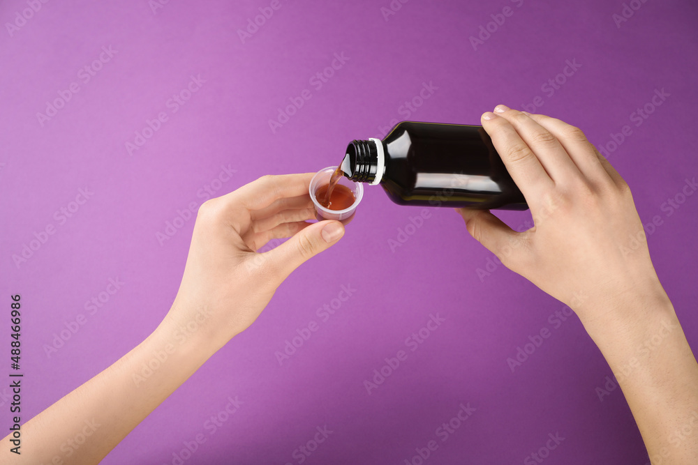 Woman pouring cough syrup into measuring cup on purple background, closeup