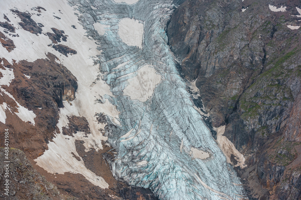 Atmospheric mountain landscape with long vertical glacier tongue with cracks among rocks. Awesome aerial view to large glacier with icefall. Nature texture of mountain glacier with fissures close up.