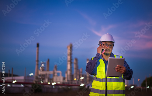 Engineer wearing safety uniform and helmet looking detail tablet on hand with oil refinery factory at background. 