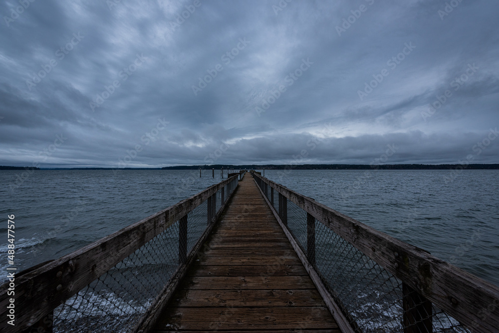 Long wooden pier stretching into the horizon over water on cloudy overcast blue hour morning