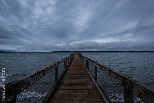 Fototapeta Naklejka Na Ścianę i Meble -  Long wooden pier stretching into the horizon over water on cloudy overcast blue hour morning