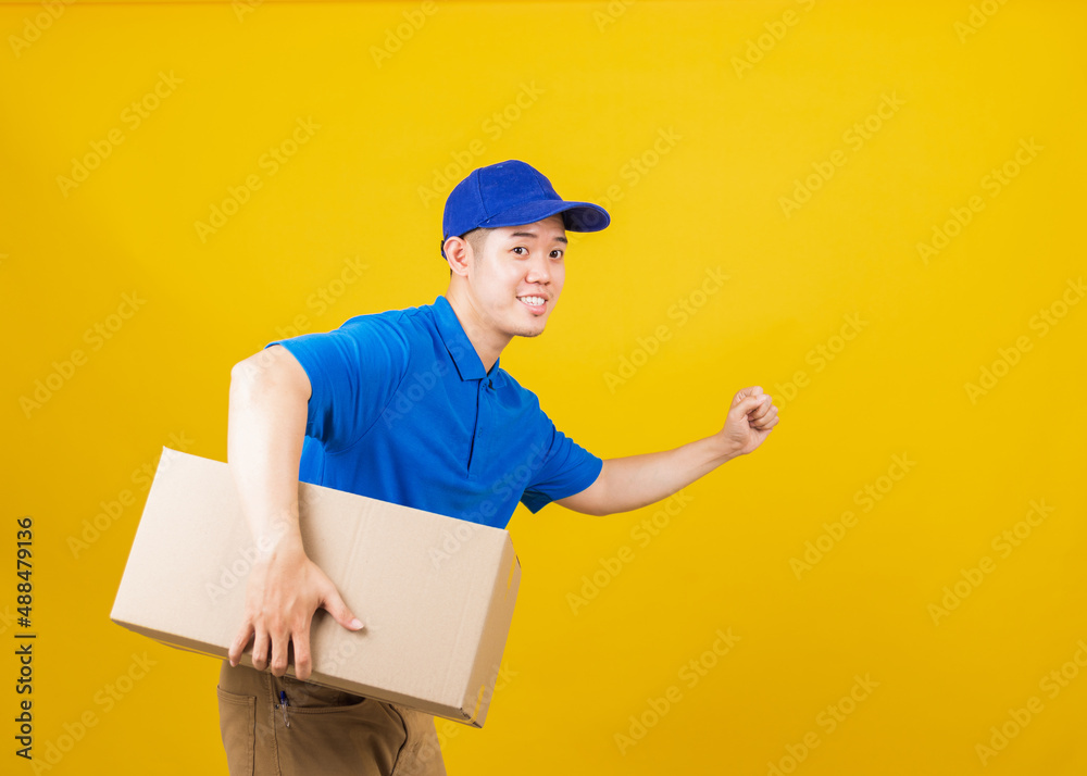 Portrait excited delivery happy man logistic standing smile wearing blue t-shirt and cap uniform holding parcel box and running looking to camera, studio shot isolated on yellow background, side view Portrait excited delivery happy man logistic standing smile wearing blue t-shirt and cap uniform holding parcel box and running looking to camera, studio shot isolated on yellow background, side view