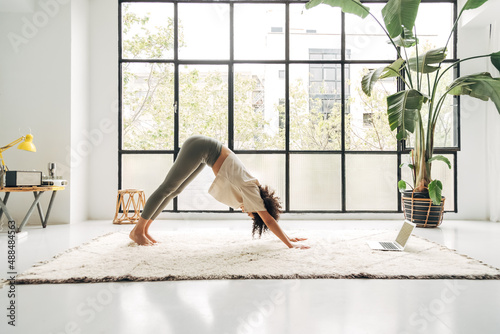 Young multiracial woman doing downward facing dog yoga pose at home watching online class using laptop. Copy space.